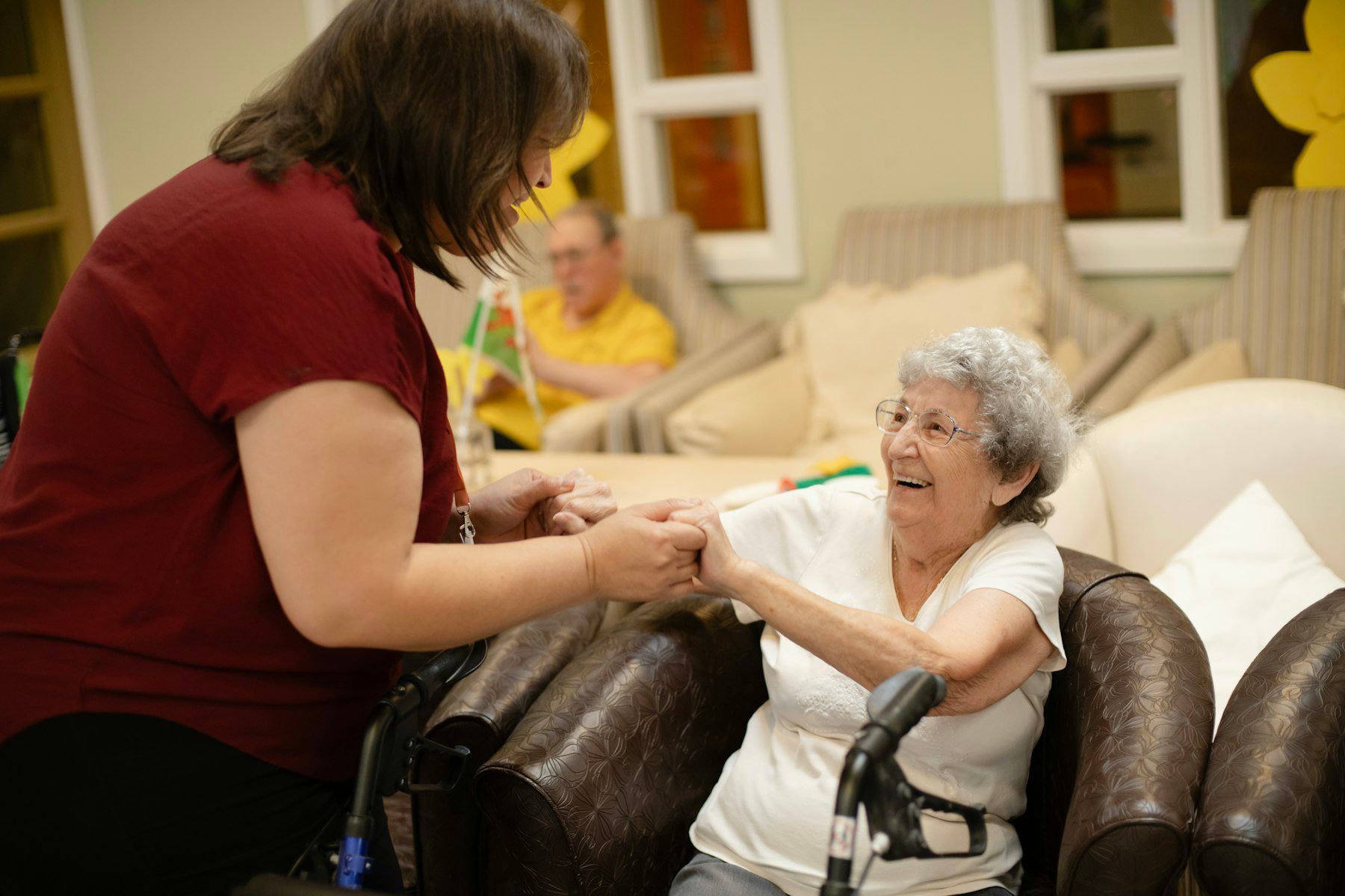 A caregiver holds the hands of a smiling older woman using a walker in her living room