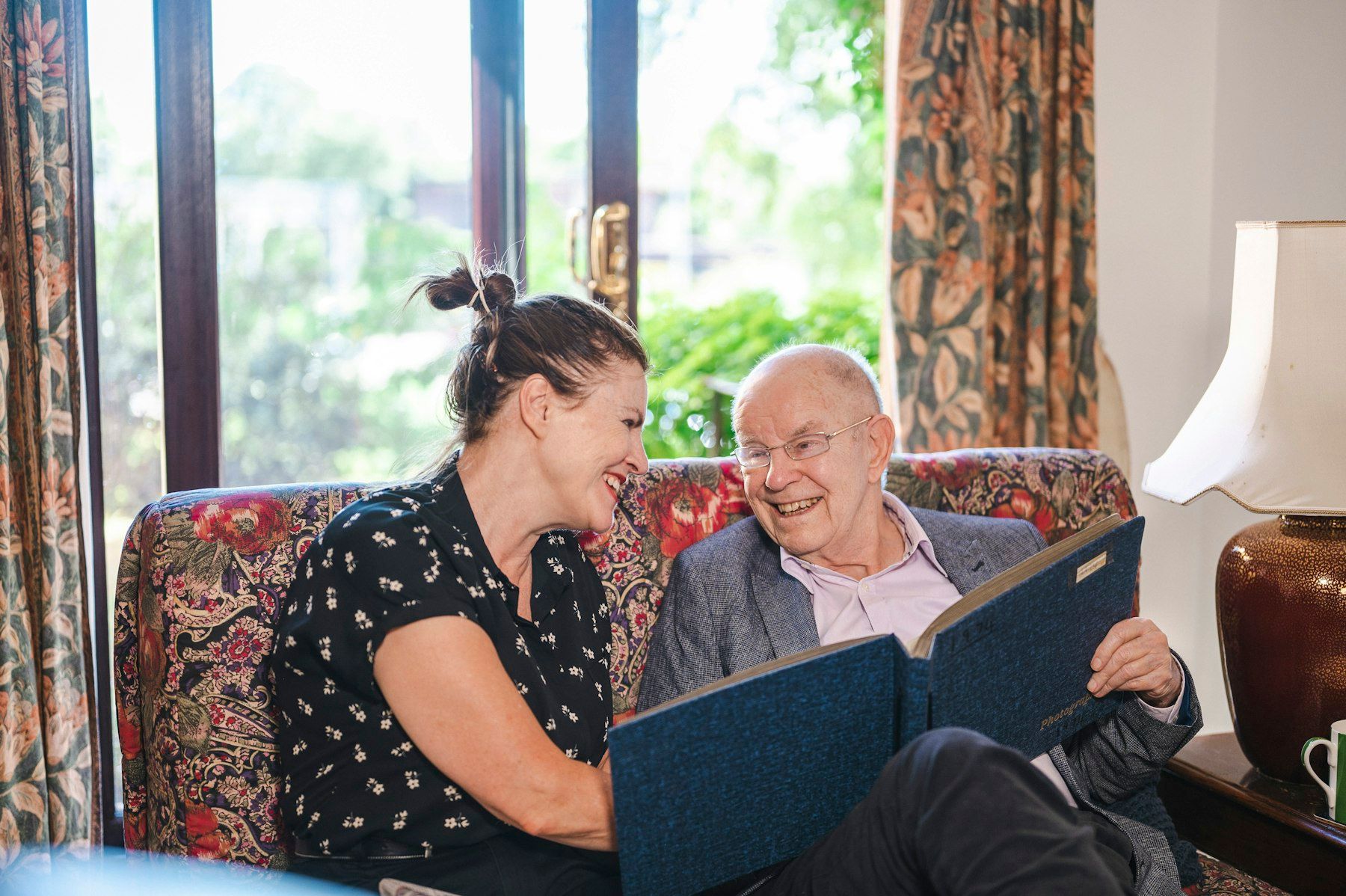 A woman and an older man laugh over a photo album on the sofa