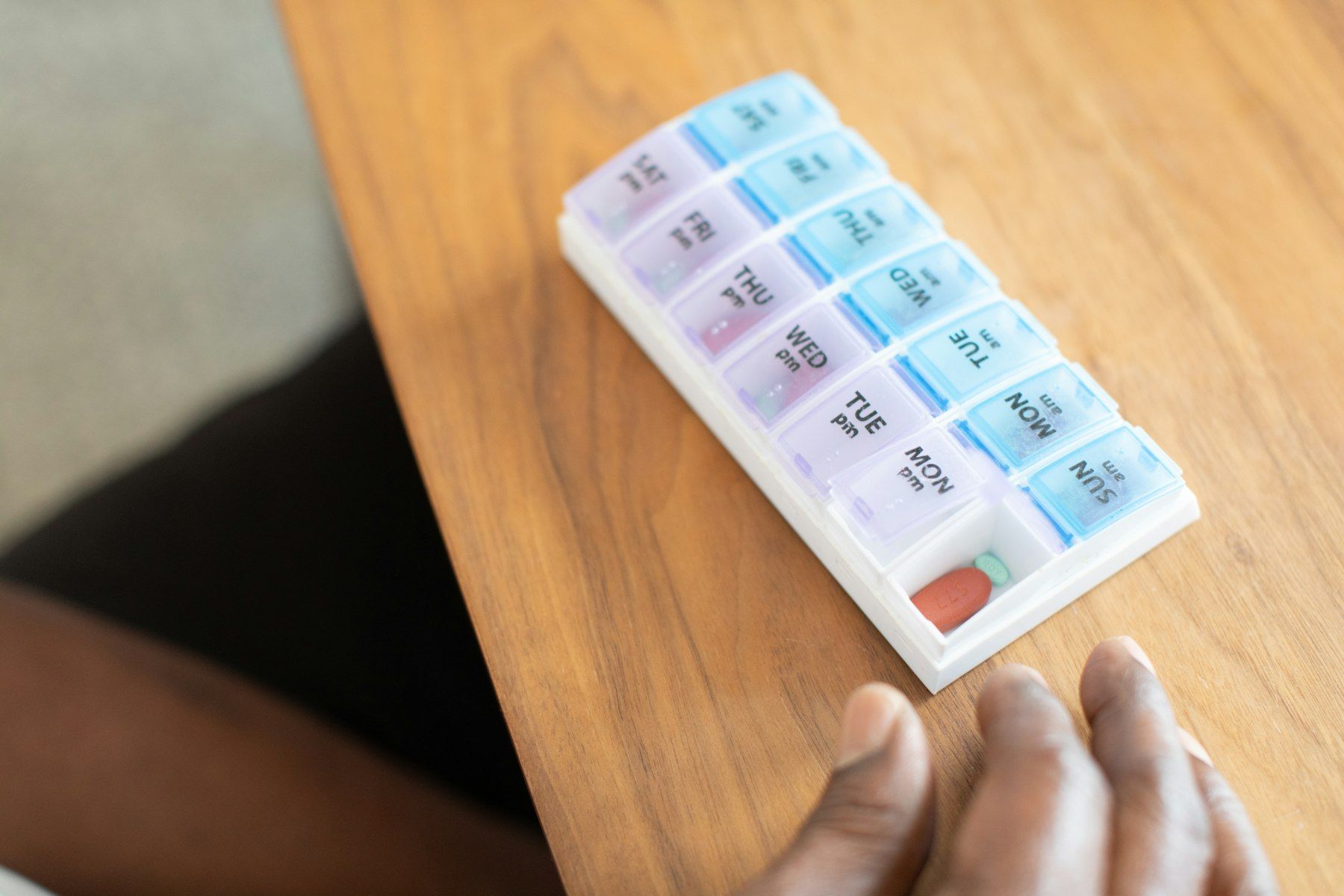 A hand reaches for a weekly pill organiser on a wooden table