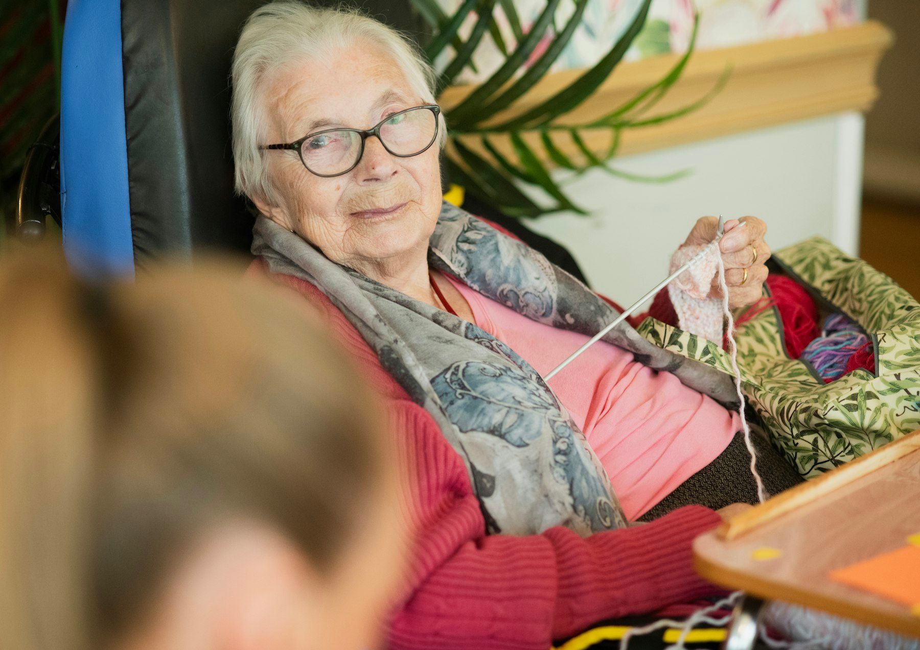 An older woman smiles from her chair, knitting on her lap